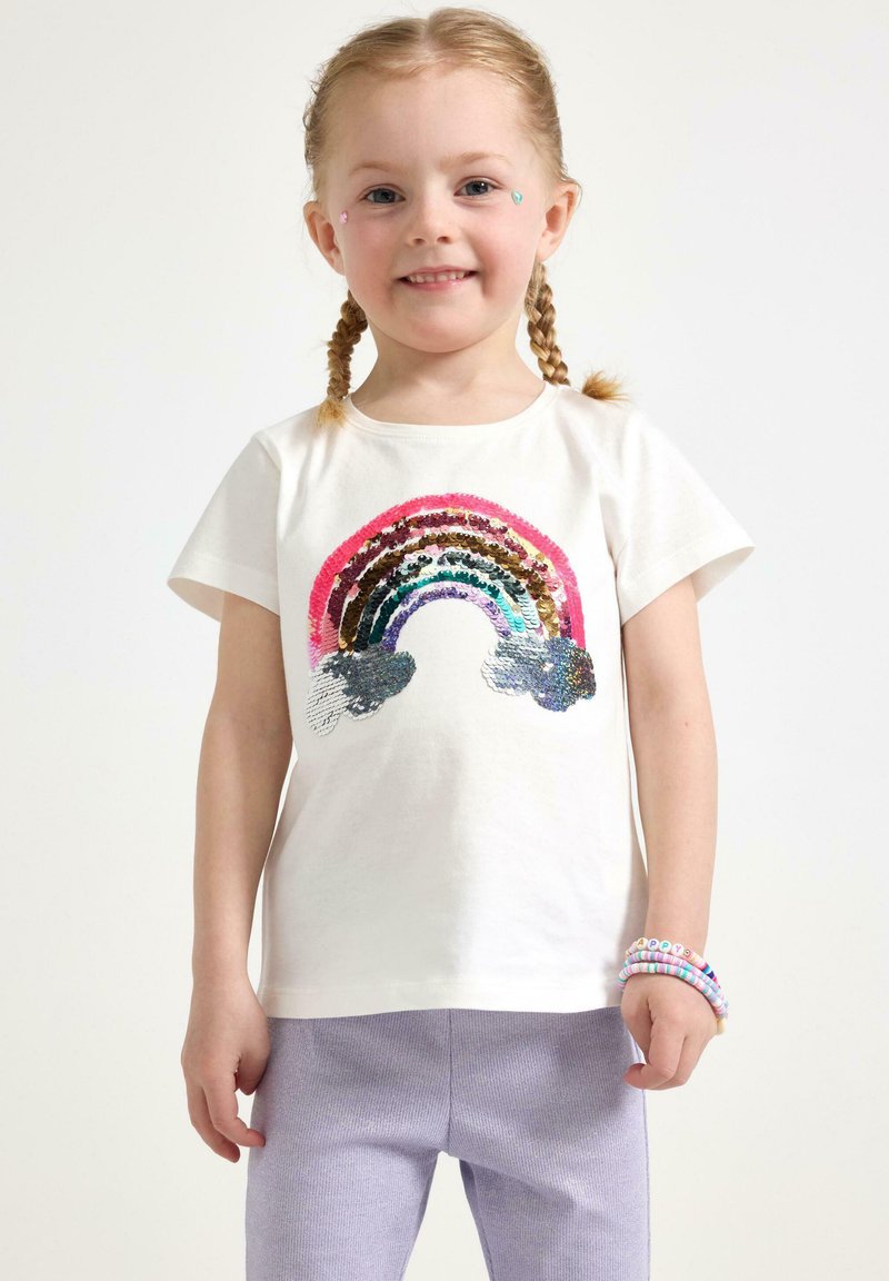 Vit bomulls-T-shirt med en flerfärgad paljettregnbågsdesign. Korta ärmar, bärs med lila stickade byxor och färgglada pärlarmband.
