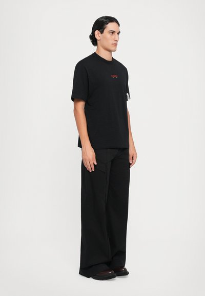 Schwarzes Baumwoll-T-Shirt mit kleinem rotem Logo, kombiniert mit lockeren schwarzen Cargo-Hosen. Gummisohle Schuhe vervollständigen das Outfit. Seitenansicht.