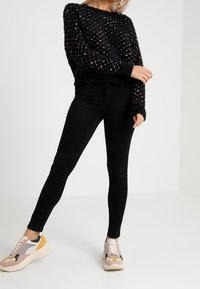 Czarny dziergany sweter z wielokolorowymi kropkami, zestawiony z dopasowanymi czarnymi jeansami i masywnymi sneakersami z różowymi, szarymi i żółtymi akcentami.