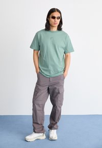 T-shirt de algodão verde-claro com um pequeno logotipo, combinado com calças cargo cinzas largas. O visual é complementado com tênis brancos e óculos de sol.