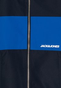 Mėlyna ir juoda striukė su pilnu užtrauktuku, turinti dvi horizontalias juosteles ir prekės ženklo pavadinimą "JACK & JONES" baltomis raidėmis.