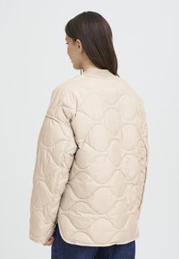 Veste matelassée beige avec un col rond, une surface texturée et une coupe ample. Présente un motif subtil en vague et des manches longues.