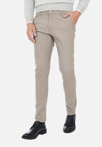Pantaloni beige slim-fit in misto cotone, con chiusura a zip, tasche frontali e design pulito. Indossati con scarpe nere stringate.