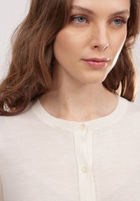 Jasno beżowy kardigan z okrągłym dekoltem, z zapięciem na guziki i prążkowanymi krawędziami. Miękka tekstura, subtelny połysk, minimalistyczny design.