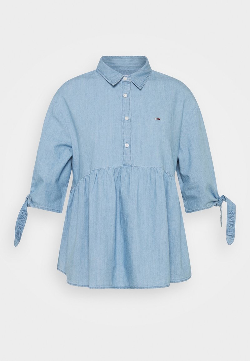 Blouse en denim bleu clair avec trois boutons, col, manches mi-longues nouées aux extrémités, et un petit logo sur la poitrine gauche.