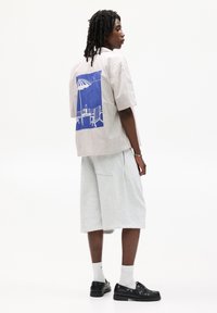 Camicia a maniche corte a righe color beige chiaro con stampa grafica blu sul retro; abbinata a pantaloni corti grigi e scarpe nere, si trova davanti a uno sfondo neutro.