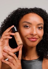 Donna con capelli ricci tiene una bottiglia di fondotinta del marchio KIKO vicino alla guancia, sorridendo dolcemente su uno sfondo neutro.