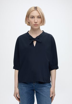 Blonde woman with short hair wearing a black top with a bow tie neckline and blue jeans, standing against a plain white background.