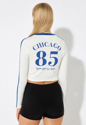 Long-sleeve, cropped white shirt with blue accents, featuring "CHICAGO 85" and "United State of America" printed on the back. Black shorts below.