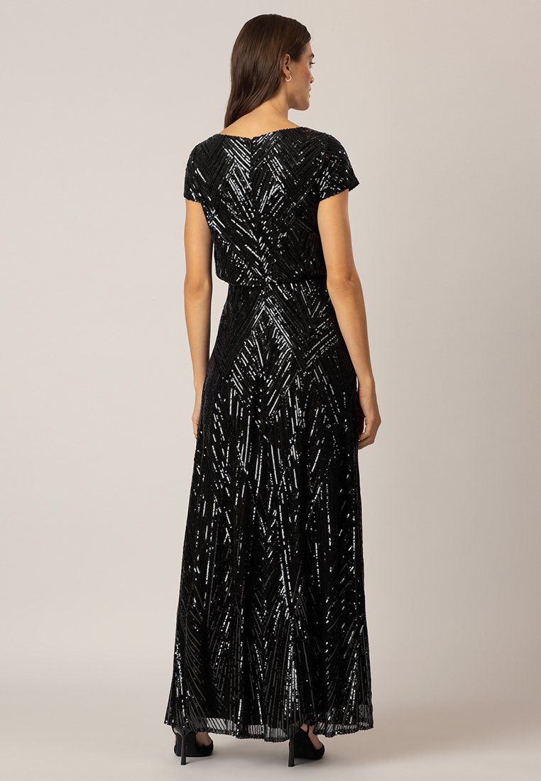 Femme portant une robe à sequins noire, longue jusqu'au sol, avec des manches courtes, se tenant de dos, mettant en valeur le motif en chevron texturé de la robe.