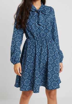 Kvinde iført en blå langærmet kjole med et blomstret geometrisk mønster og en bundet sløjfe ved kraven, stående foran en ensfarvet baggrund.