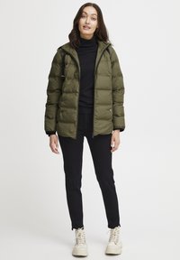 Olivovo zelená zateplená bunda so zapínaním na zips, so kapucňou, elastickými manžetami a bočnými vreckami, nosená cez čierny rolák a čierne nohavice.