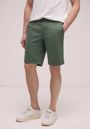 Mann trägt olivgrüne, knielange Shorts mit Taschen und weiße Sneakers, steht vor einem schlichten Hintergrund.