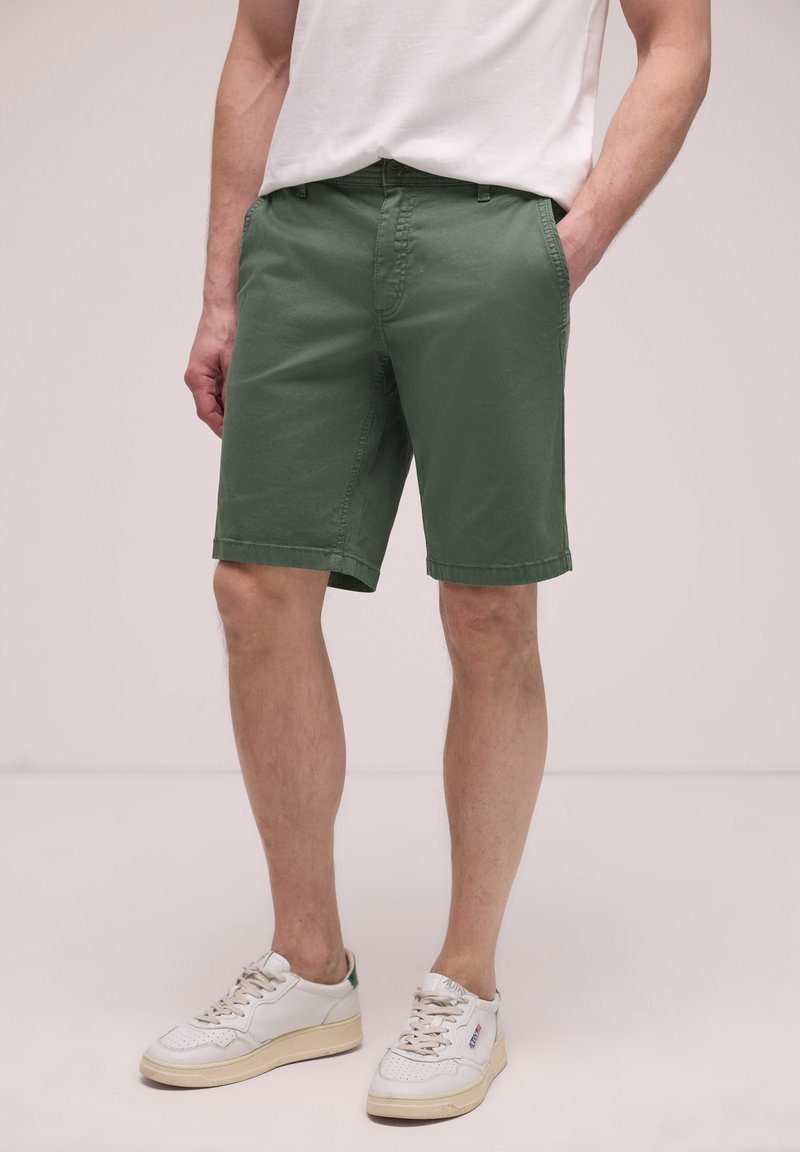 Homme portant un short longueur genou vert olive avec des poches et des baskets blanches, debout devant un fond uni.