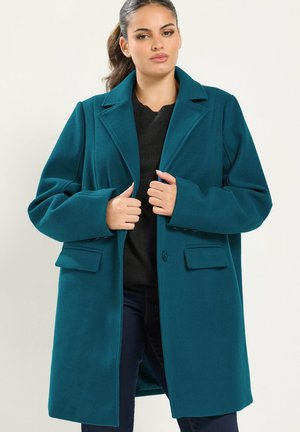 Femme aux cheveux foncés en queue de cheval, portant un long manteau bleu sarcelle foncé par-dessus un haut noir et un pantalon foncé, tenant les revers du manteau avec les deux mains.