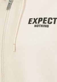Cream zip-up hoodie with a textured fabric. Black text "EXPECT NOTHING" prominently featured on the chest. Zipper detail visible.