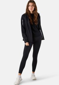 Zwarte waterdichte jas met een ritszak, gecombineerd met zwarte leggings en witte sportschoenen. Gladde textuur, atletische pasvorm.