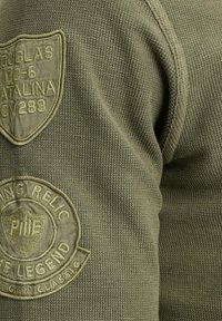 Olivgrüner Strickpullover mit strukturiertem Stoff, einem runden Logostick und einem Emblempatch auf der Schulter, das komplizierte Stickerei zeigt.