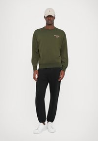 Hanorac verde închis cu manșete texturate, asortat cu pantaloni sport negri și adidași albi. Modelul poartă de asemenea o șapcă bej cu un logo.