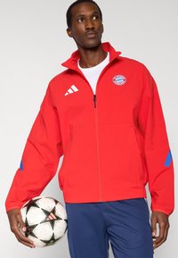 Red zip-up jacket with blue accents, featuring a logo and white stripes on the shoulders. Holding a patterned soccer ball.