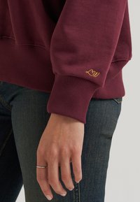 Gros plan d'une personne portant un jean bleu foncé et un sweat bordeaux avec une broderie jaune "LW" sur le poignet et une bague argentée au doigt.