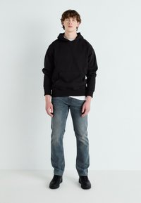 Felpa nera con cappuccio e tasca a marsupio frontale, abbinata a jeans blu scoloriti e scarpe da ginnastica nere, su uno sfondo chiaro.