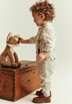 Young child with curly hair feeds a stuffed fox sitting on a wooden chest in a neutral-toned setting.