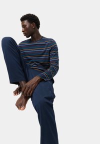 Camicia a maniche lunghe a righe navy con sottili linee multicolori, abbinata a pantaloni navy scuri; modello seduto, che tiene un piede.