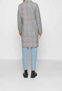 Grau-schwarz karierten Mantel mit taillierter Passform, ausgestattet mit langen Ärmeln und einem einzelnen Rückenschlitz, kombiniert mit hellblauen Jeans und schwarzen Schuhen.