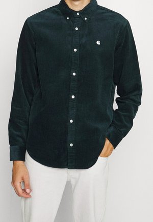 Mężczyzna w ciemnozielonej koszuli sztruksowej zapinanej na guziki z białymi guzikami, logo na piersi, i białych spodniach, ręka w kieszeni.