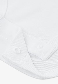 Λευκό βαμβακερό ύφασμα με υφή στην επιφάνεια. Διαθέτει γιακά και κλείσιμο με κουμπιά, παρουσιάζοντας λεία φινίρισμα και διακριτικές λεπτομέρειες ραφής.