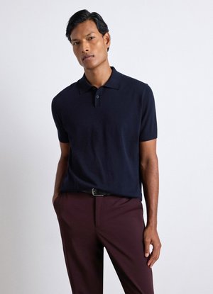 Mann mit kurzen schwarzen Haaren, der ein dunkelblaues Polohemd und dunkelburgunderfarbene Hose trägt und mit einer Hand in der Tasche vor weißem Hintergrund steht.