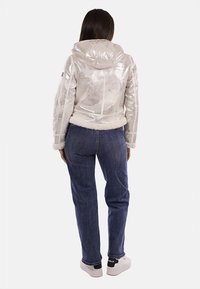 Giacca argentata, lucida, con zip e cappuccio, foderata in pelliccia sintetica; abbinata a jeans blu e sneakers bianche con dettagli neri.