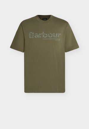 Jeune homme portant un t-shirt Barbour vert olive, un jean bleu, des baskets blanches, tenant un sac fourre-tout beige, debout devant un fond uni.