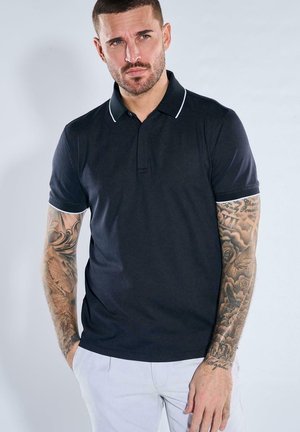 Man with short hair and full arm tattoos wearing a black polo shirt and light-colored pants, standing with hands partially in pockets.