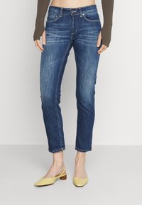 Pantalones vaqueros de mezclilla azul oscuro con corte ajustado, cinco bolsillos y costuras de contraste. Combinados con zapatos de tacón amarillos, con punta afilada.