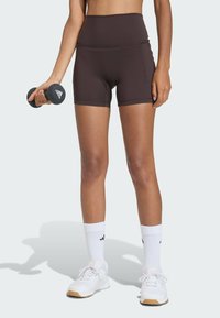 Hnedé šortky s vysokým pásom vyrobené z elastickej látky, kombinované s bielymi športovými ponožkami a topánkami. Drží čiernu činelu. Viditeľné logo Adidas.