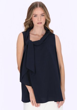 Giovane donna che indossa un top senza maniche blu navy con dettaglio asimmetrico di tessuto intorno allo scollo e pantaloni bianchi su sfondo neutro.