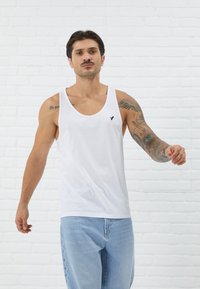 White cotton tank top with wide armholes and a round neckline, featuring a small black logo on the chest. Worn with light blue jeans.