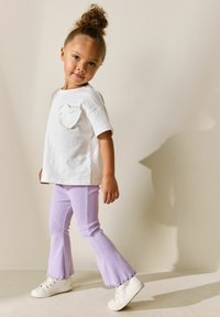 Biele tričko s krátkym rukávom a vreckom v tvare srdca, doplnené svetlofialovými rozšírenými nohavicami s rebrovanou textúrou a bielymi teniskami. Jednoduchý, hravý dizajn.