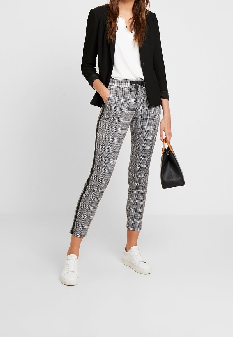 Femme portant un blazer noir, un haut blanc, un pantalon à carreaux avec bande latérale, des baskets blanches, tenant un sac à main noir avec des poignées en bois.