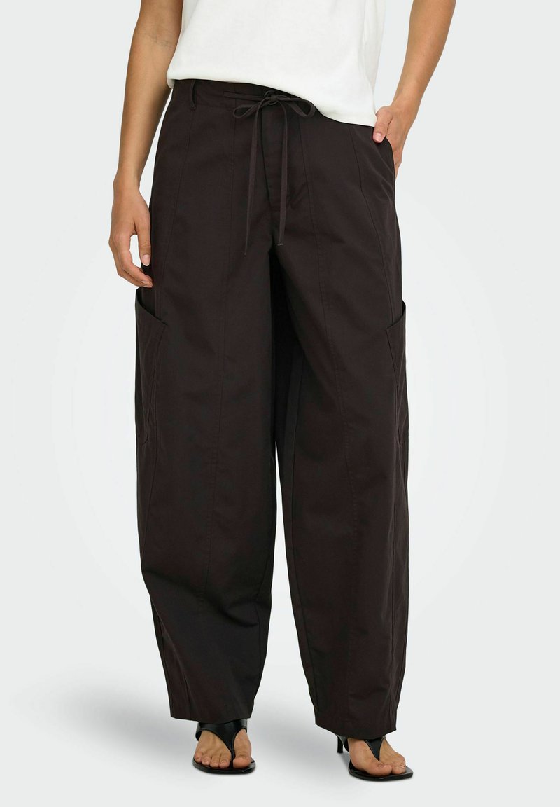 Personne portant un pantalon noir ample avec de grandes poches latérales, un haut blanc et des sandales à talons noires ouvertes à l'avant, debout sur un fond uni.