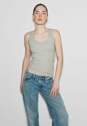 Naine, kellel on tagasikeeratud juuksed, kannab helerohelist lainelise äärega toppi ja siniseid teksaseid, seisab tavalise valge tausta ees.