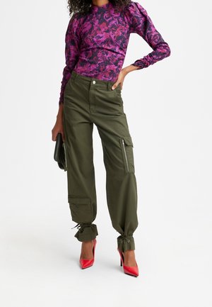 Frau trägt ein langärmliges Oberteil mit lila Muster, olivgrüne Cargo-Hose mit Reißverschlusstaschen, rote High Heels und hält eine dunkle Clutch-Tasche.