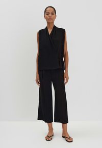 Haut noir sans manches à nouer sur le côté, associé à un pantalon noir à jambes larges et des sandales plates noires. Tissu lisse avec un design minimal.