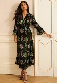 Schwarzes, floralbedrucktes Maxikleid mit V-Ausschnitt, Rüschensäumen und kunstvollen Blattformen in Lila, Grün und Blau. Der durchsichtige Stoff fällt elegant.