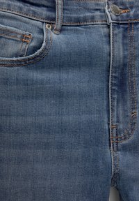 Gros plan sur un jean en denim bleu montrant la poche avant, la petite poche, les passants de ceinture, le bouton en métal et les détails de couture orange.