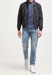 Černá bomber bunda s červeným akcentem, modrá košile s knoflíky, světle modré džíny s efektem opotřebení a bílé tenisky. Model stojí, ruce jsou viditelné.