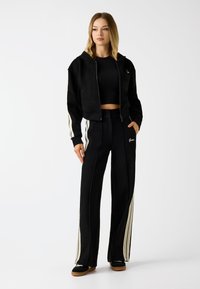 Schwarzer, kurz geschnittener Hoodie mit Reißverschluss und Logo, kombiniert mit hochgeschnittenen schwarzen Hosen mit weißen seitlichen Streifen und gesticktem Branding. Lässiges Design.