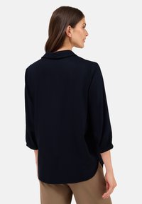 Frau mit langen braunen Haaren, die eine schwarze Bluse mit langen Ärmeln und beige Hosen trägt, von hinten vor einem schlichten Hintergrund gezeigt.